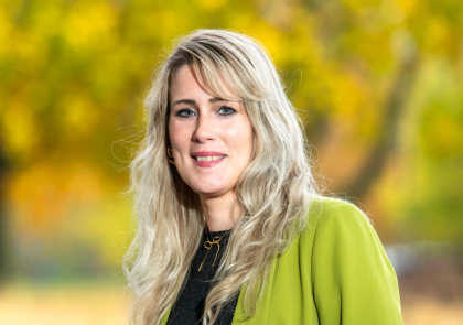 Foto van vrouw met blond haar en groen jasje tegen een herfstige achtergrond. 